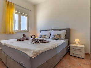 Uma cama bem arrumada com um edredom branco e duas almofadas, uma mesa de cabeceira com abajur e uma janela com cortinas amarelas.