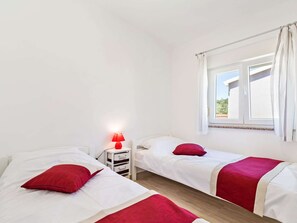 A twin room with two beds, each with a red pillow and a white bedspread. There is a nightstand with a red lamp and storage drawers. A window with white curtains provides a view of the outside. - Stylish Holiday Home in Banjole With Swimming Pool (Medulin)