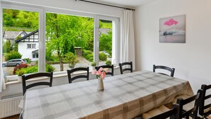 A dining area with a table set for four, large windows with curtains, a radiator, and a painting on the wall. - Ferienhaus Bergfreiheit (Winterberg)