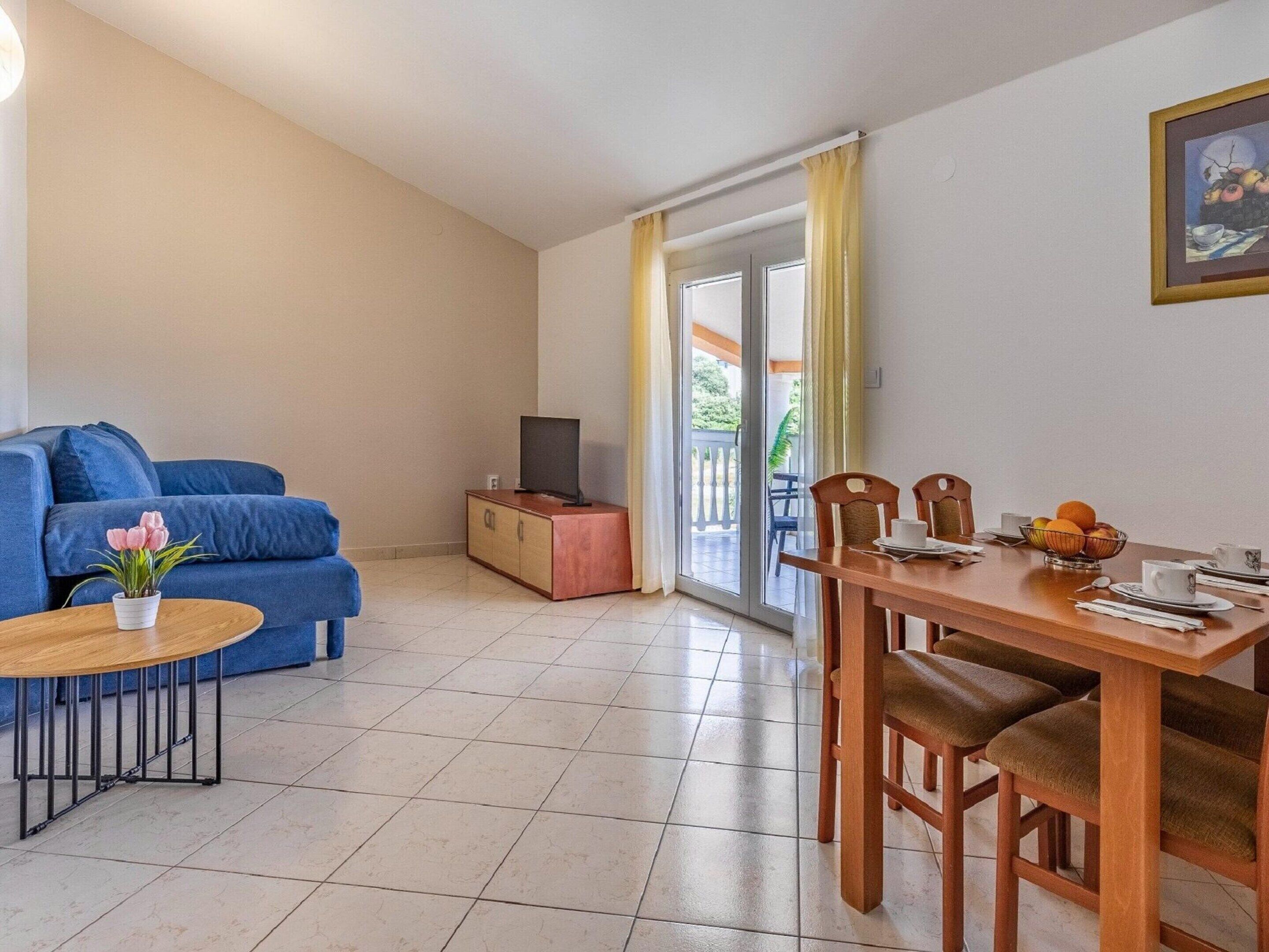 Un salon avec un canapé bleu, une petite table sur laquelle est posée une plante, un téléviseur sur un meuble en bois, et un coin repas avec une table dressée pour deux personnes.