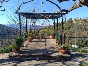 Un'area esterna con pavimentazione in pietra, un pergolato in metallo, piante in vaso e vista su un paesaggio montuoso.