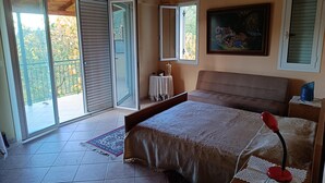 A bedroom with a bed, a bedside table, a nightstand, a lamp, a painting, and a sliding glass door leading to a balcony. - Hillview Holiday Home in Kalavrouza (Nafpaktos)