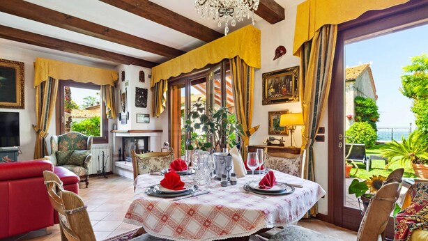 A dining area with a table set for four, a red sofa, and a view of the garden and sea. - Belvilla by OYO Villa Pratolina Agli Alberoni (Venice)