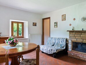 A cozy living room with a fireplace, a dining table with chairs, and a window with a view of greenery. - Apartment in Fosciandora With Pool (Fosciandora)