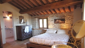 Ein Schlafzimmer mit Steinwand, Holzbalken, einem Bett mit gemustertem Überwurf, einem Schminktisch, einem Spiegel und einem kleinen Tisch mit Stuhl.