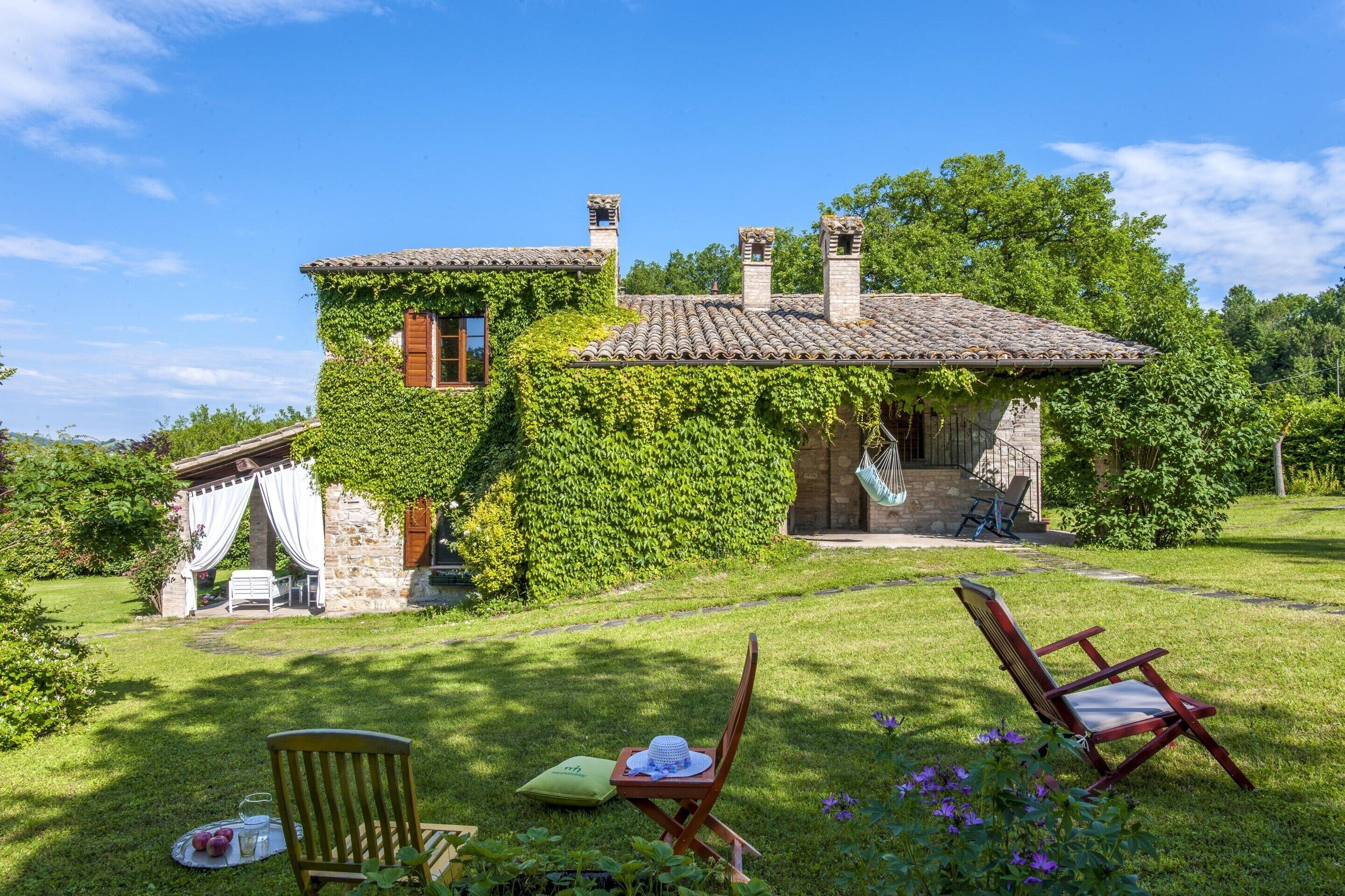 Une maison en pierre aux murs recouverts de lierre, avec un toit de tuiles et un petit porche agrémenté d’une chaise et d’une table.