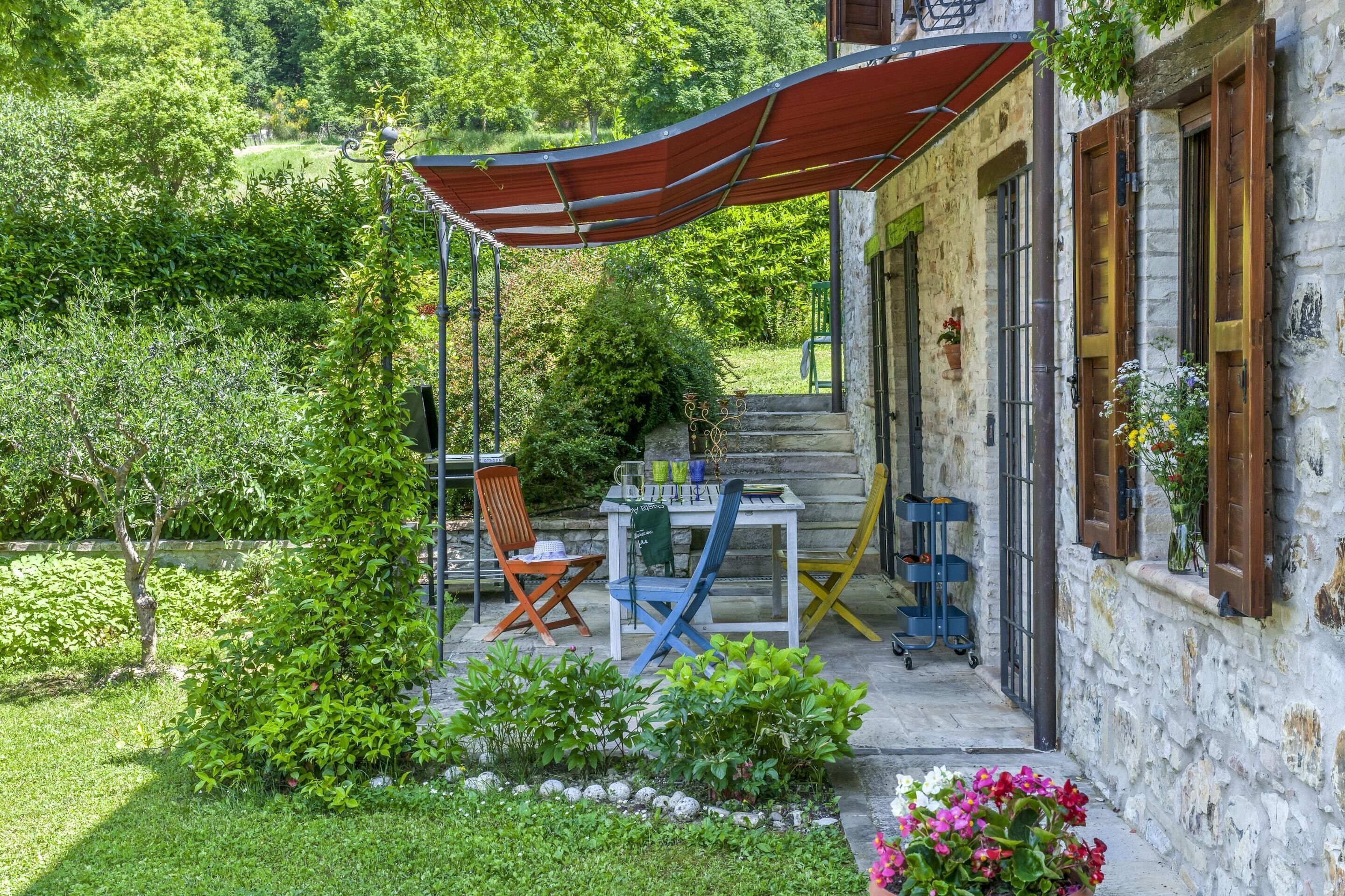 A stone building with a covered outdoor seating area, a table with chairs, and a garden with various plants and flowers.