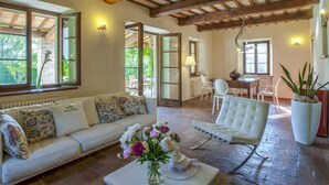 A spacious living room with a sofa, armchair, and dining table. There is a large window providing natural light and a view of the outdoors. - Marche Villa Close Culture (Fermignano)
