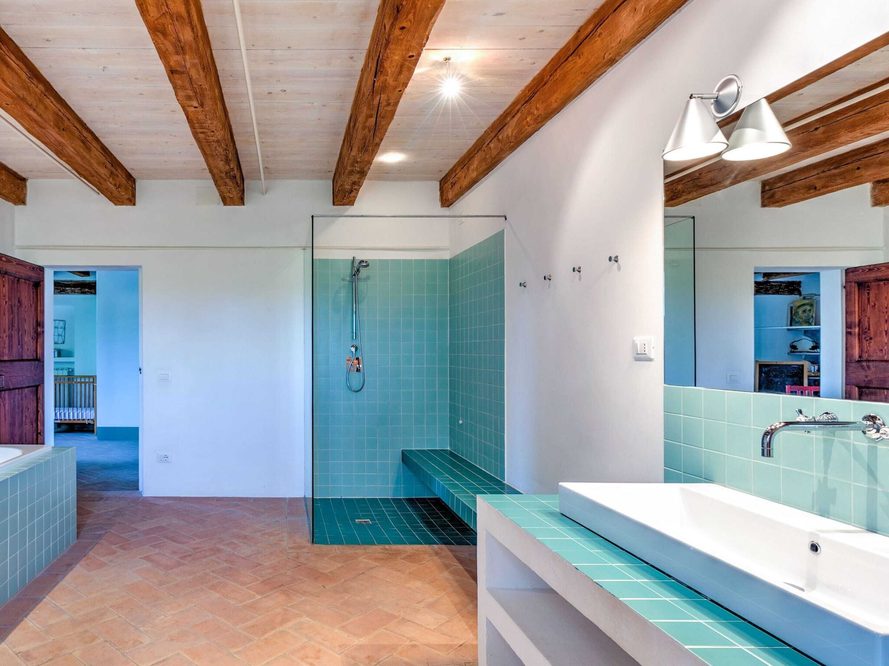 A modern bathroom with a large bathtub, a shower with blue tiles, wooden beams on the ceiling, and a mirror with a light fixture.