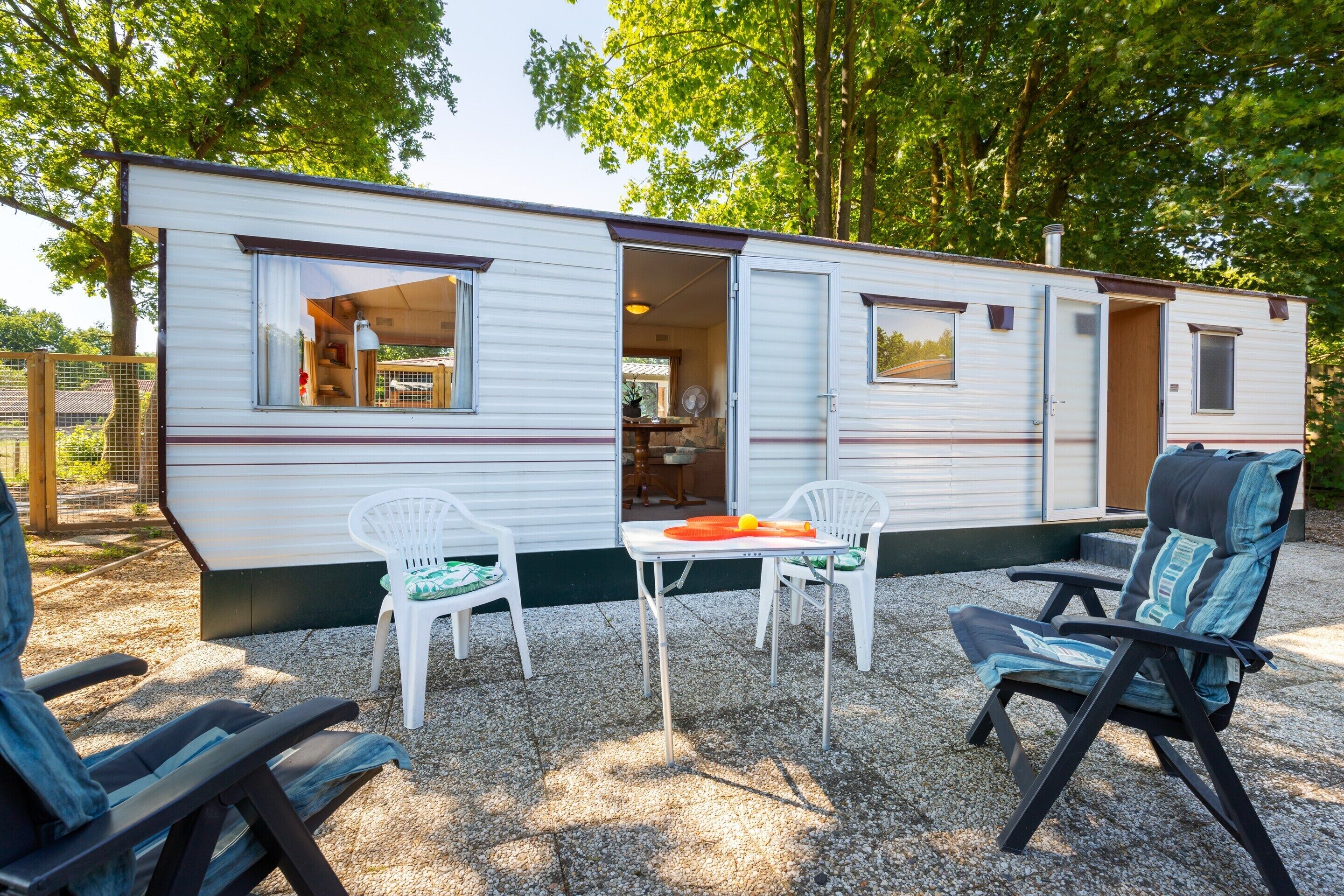 Una casa móvil con la puerta abierta que muestra una cocina, una mesa de comedor con sillas, y un porche con mobiliario exterior.