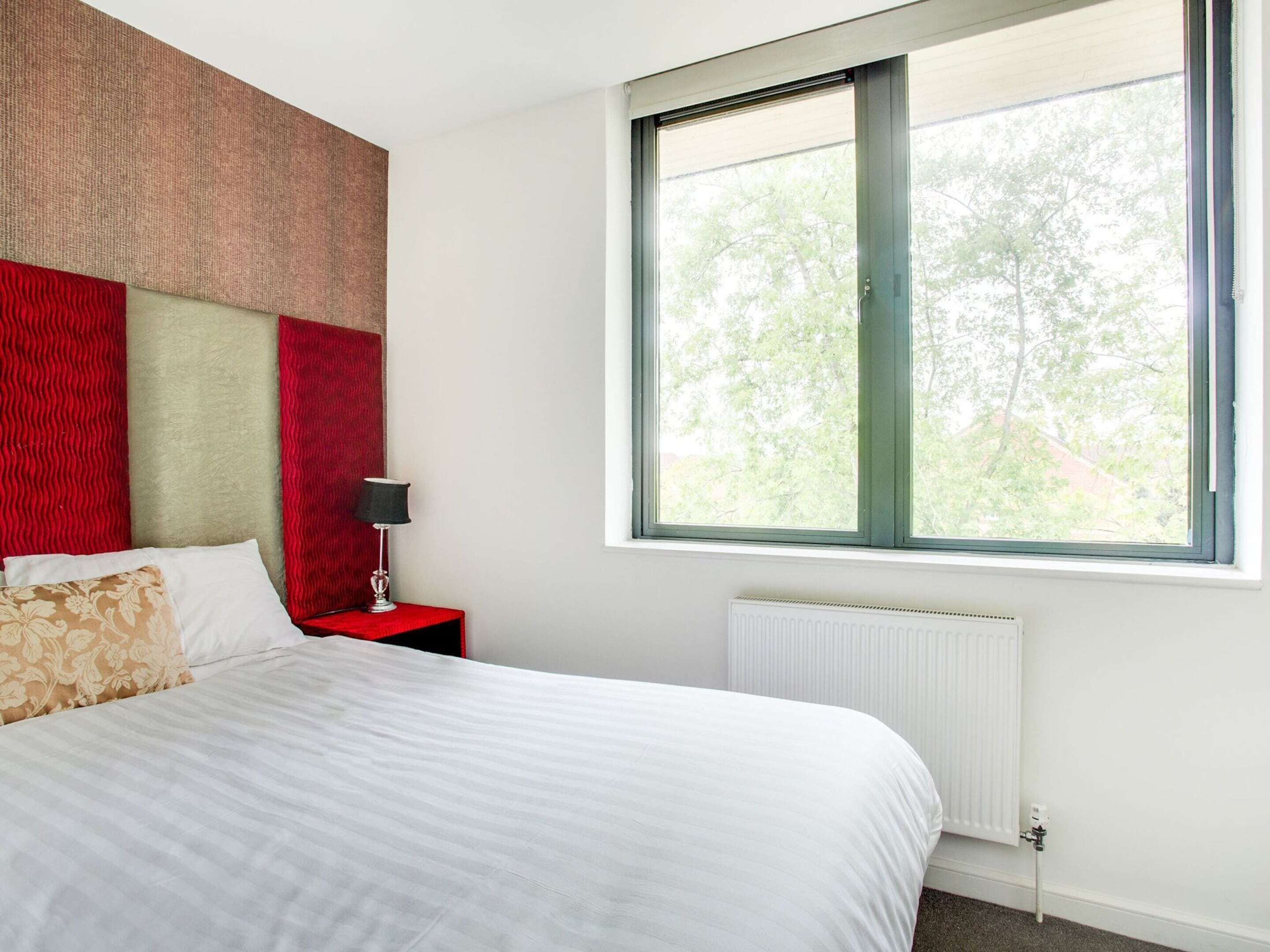 Un dormitorio con una cama grande, una mesita de noche con lámpara, un radiador y una ventana con vistas a árboles.