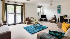 A modern living room with a sofa, armchair, and a coffee table. There is a TV and a small table with a plant. The room has a large sliding glass door leading to an outdoor area.