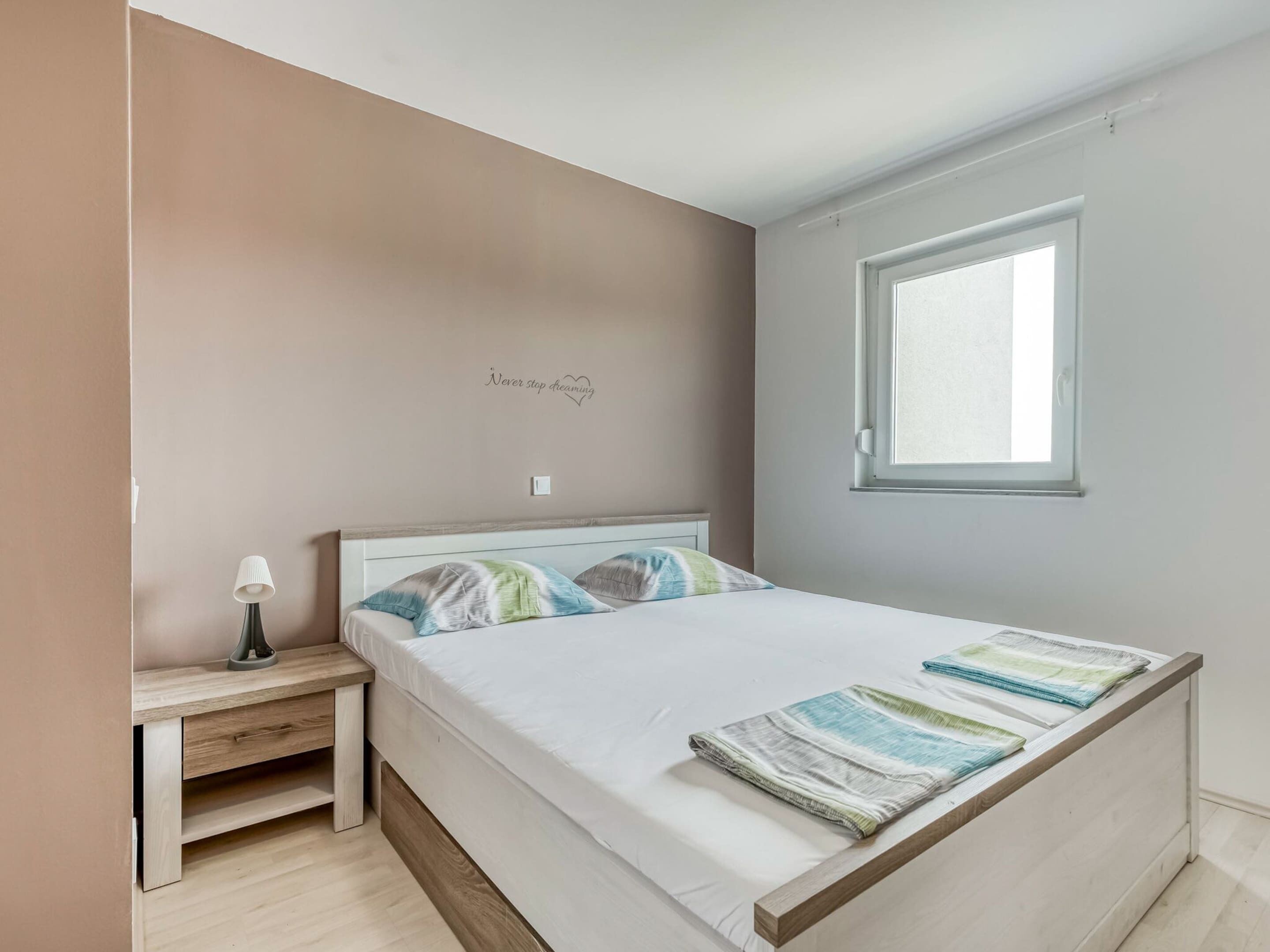 A bedroom with a bed, a nightstand, a lamp, a window, and a motivational quote on the wall.