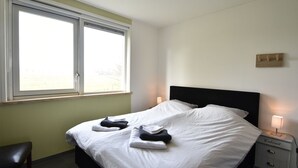 A neatly made bed with white linens and towels, a bedside table with a lamp, and a window with blinds. - Lovely Cottage in the Middle of Nature (Keijenborg)