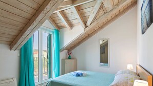 Um quarto com teto inclinado, uma cama com colcha verde, uma mesa de cabeceira com abajur e uma janela com cortinas.