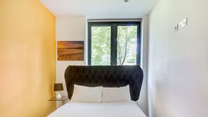 Ein Schlafzimmer mit einem Bett, einem Nachttisch, einer Lampe, einem Gemälde und einem Fenster mit Blick auf Bäume.