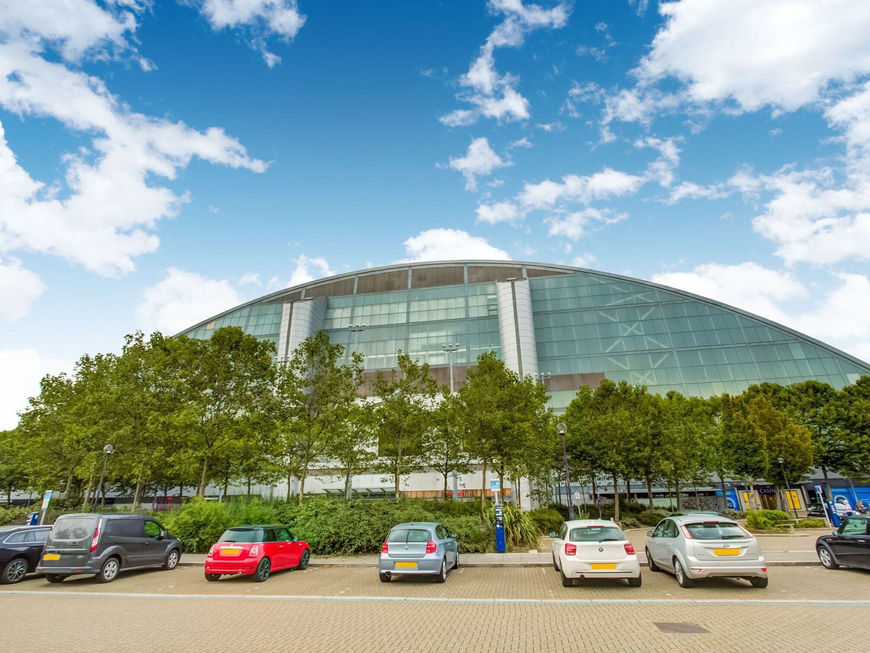 A modern building with a curved glass facade, surrounded by parked cars and trees.