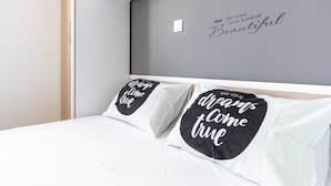 Quarto com cama, duas almofadas com frases motivacionais e parede com mensagem positiva.