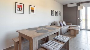 Uma sala de estar moderna com uma mesa de jantar de madeira, bancos e um sofá. Há quadros nas paredes e um aparelho de ar condicionado fixado na parede.