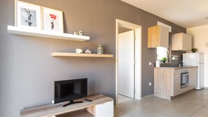 Um apartamento moderno com uma TV sobre um suporte de madeira, uma cozinha com fogão e geladeira, e uma porta que dá para outro cômodo.