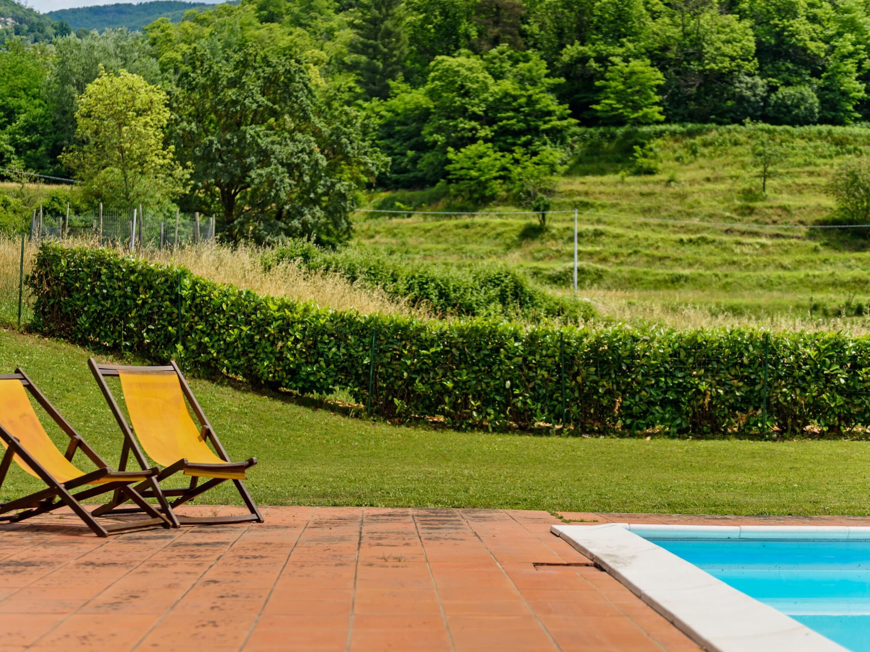 Un espace aménagé au bord de la piscine, avec une terrasse en briques, deux chaises longues jaunes et une vue sur un paysage verdoyant, avec des arbres et des collines en arrière-plan.