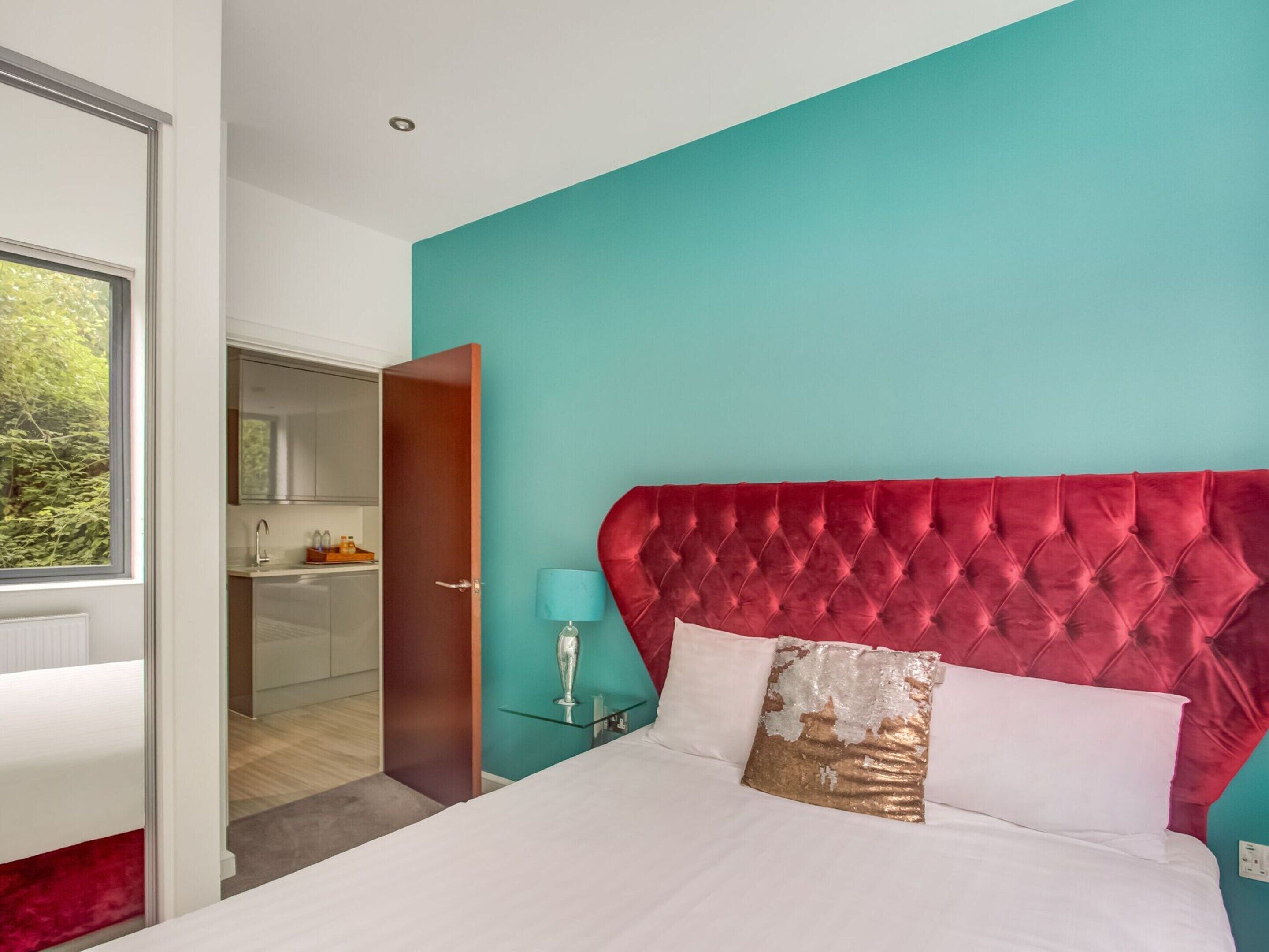 Un dormitorio con cabecera tapizada roja, pared de acento color verde azulado y edredón blanco.