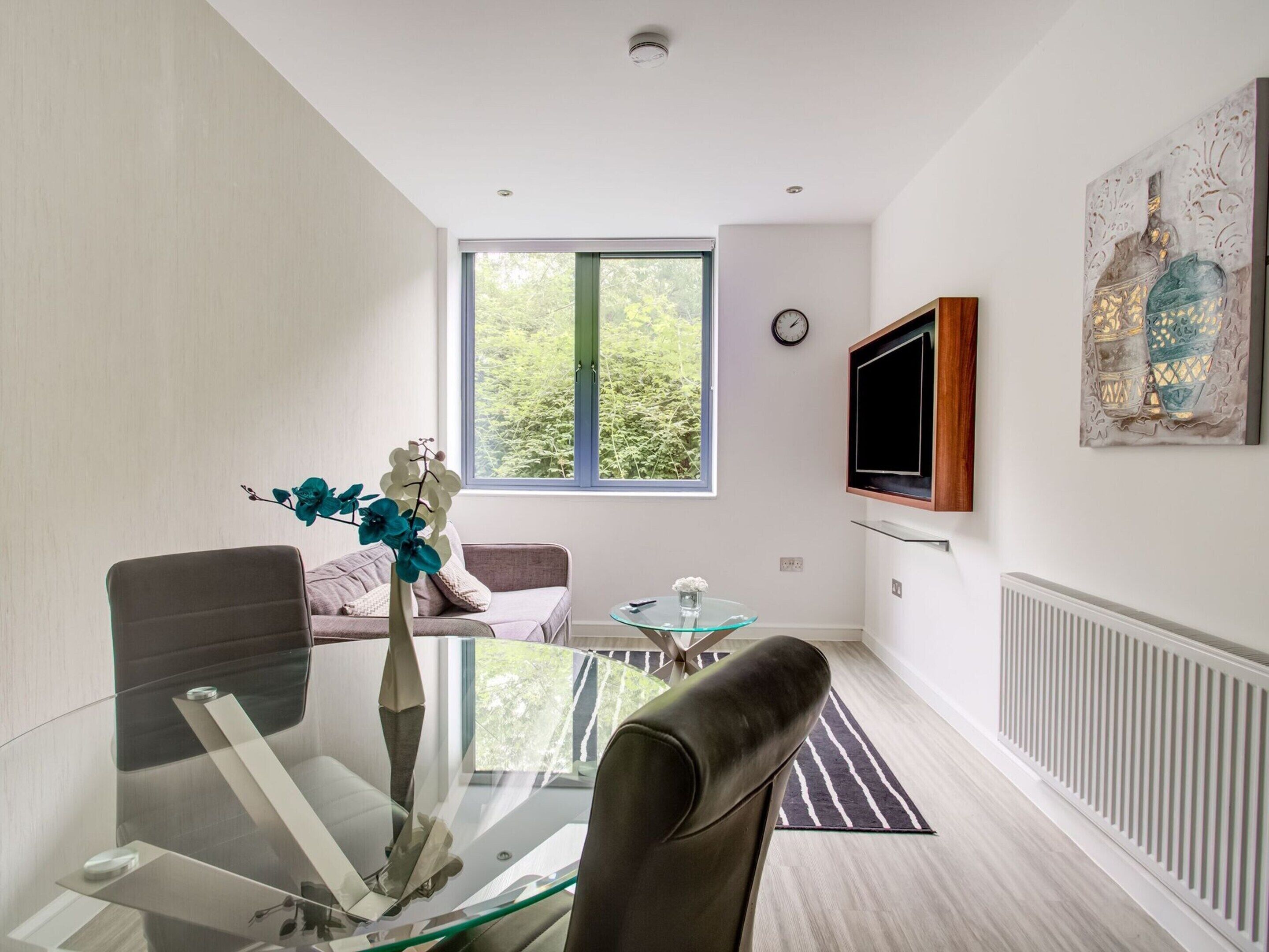 A modern living room with a glass dining table, leather sofa, and a large window offering a view of greenery.