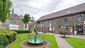 A courtyard with a playground, a brick building with a restaurant, and a church spire in the background. - Cozy Apartment in Waimes near Ovifat & Robertville Lake (Waimes)