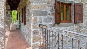 A stone building with wooden shutters, a balcony with a metal railing, and a view of greenery in the background.
