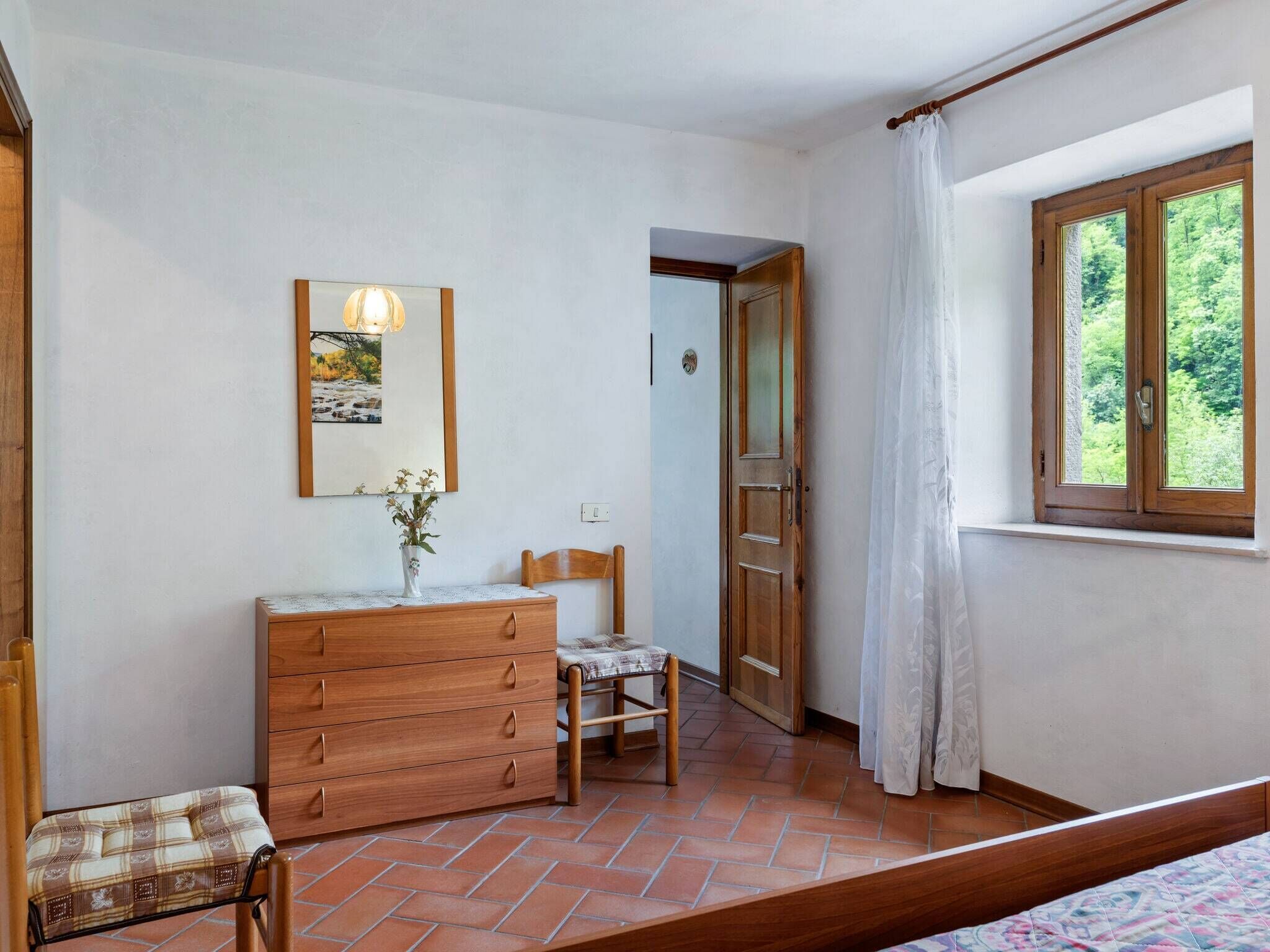 Une chambre avec une commode en bois, une chaise, un lit, une fenêtre donnant sur un paysage verdoyant et une porte menant à une autre pièce.