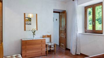 Une chambre avec une commode en bois, une chaise, un lit, une fenêtre donnant sur un paysage verdoyant et une porte menant à une autre pièce.