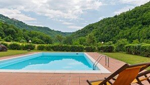 A rectangular swimming pool with a tiled deck, surrounded by lush greenery and hills.
