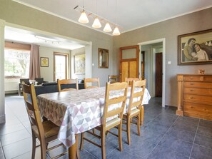 Un coin repas avec une table dressée pour quatre personnes, des chaises en bois et une nappe à motifs. Un tableau est accroché au mur et, sur le côté, on aperçoit une armoire en bois.