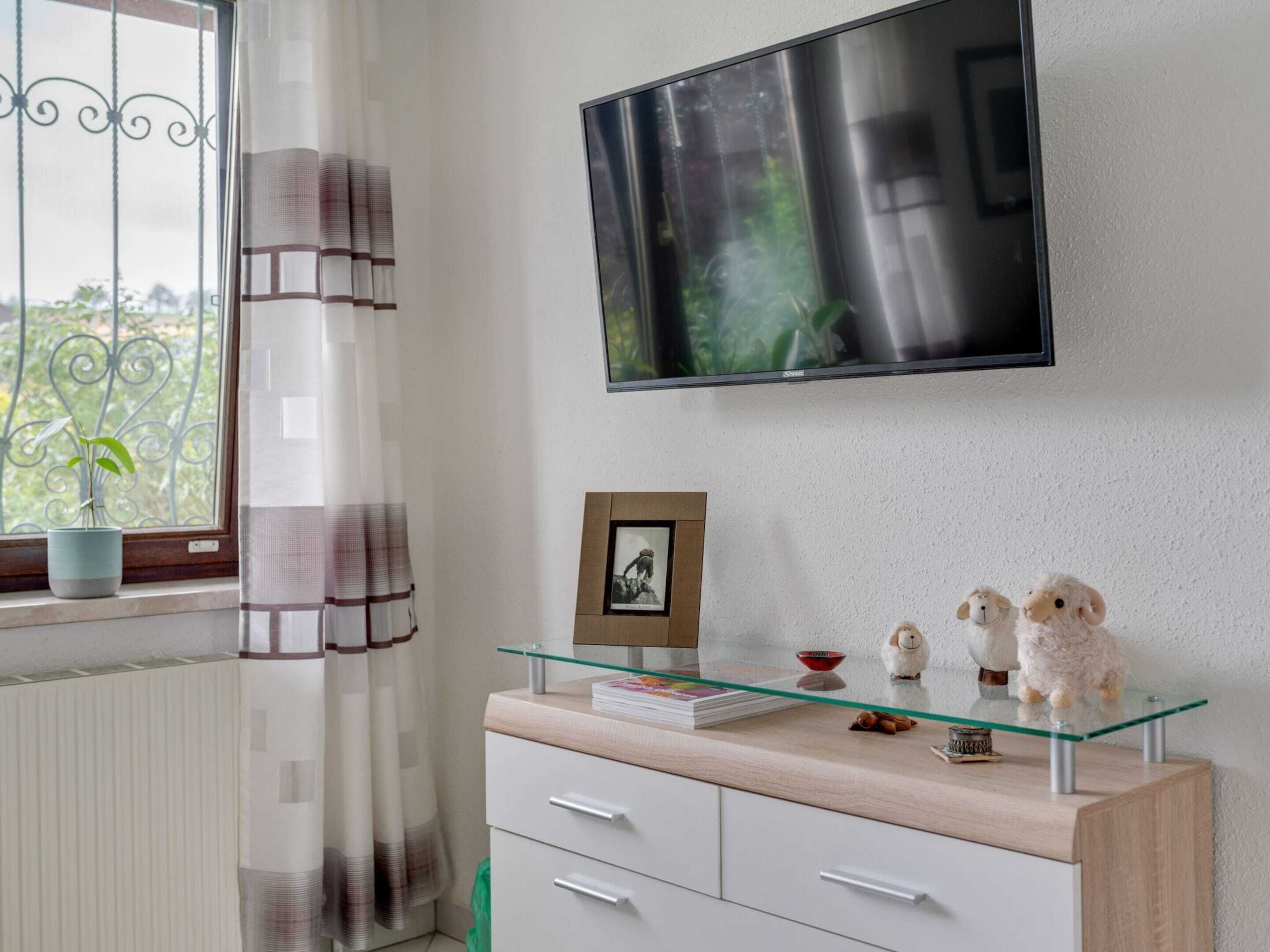 Een moderne woonkamer met een aan de muur bevestigde televisie, een salontafel met glazen blad en decoratieve objecten, een potplant bij het raam, en een witte kast met lades.
