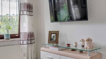 Een moderne woonkamer met een aan de muur bevestigde televisie, een salontafel met glazen blad en decoratieve objecten, een potplant bij het raam, en een witte kast met lades.