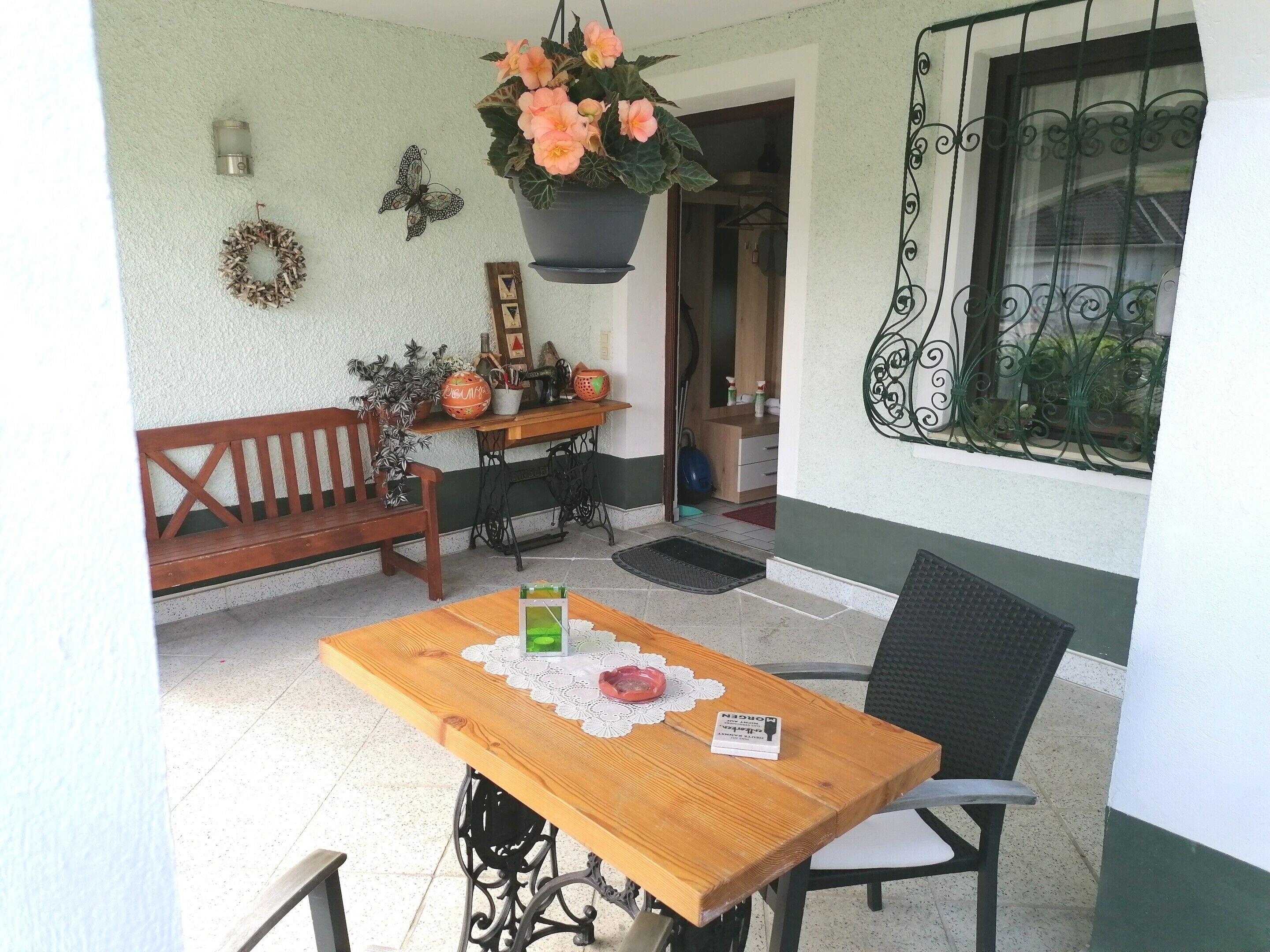A wooden table with a lace doily, a chair, a bench, a hanging flower pot, a sewing machine, and a wall-mounted light fixture.