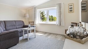 A modern living room with a grey sofa, a small white table, a lamp, a window with curtains, and a decorative tray with glass bottles. - Holiday Home in Doberan With Garden (Kuehlungsborn)