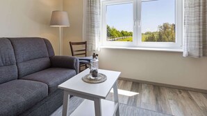 A modern living room with a grey sofa, a small white table with a decorative item, a lamp, and a window with curtains. - Holiday Home in Doberan With Garden (Kuehlungsborn)