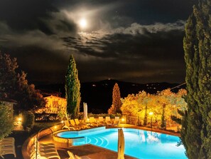 Une piscine illuminée la nuit, avec une lune en forme de croissant dans le ciel, entourée d’arbres et surplombant une chaîne de montagnes lointaine.
