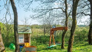 Una casita de juegos de madera rústica con techo verde, un columpio y un cobertizo pequeño en un área cubierta de césped, rodeada de árboles.
