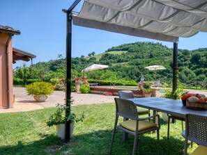 Una terrazza esterna con tavolo e sedie, circondata da piante in vaso e vista su un pendio verdeggiante.