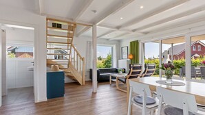 A spacious living area with a wooden staircase, a dining table with chairs, and a view of a red house through large windows. - Holiday Home Near the Baltic Sea Beach (Zierow)