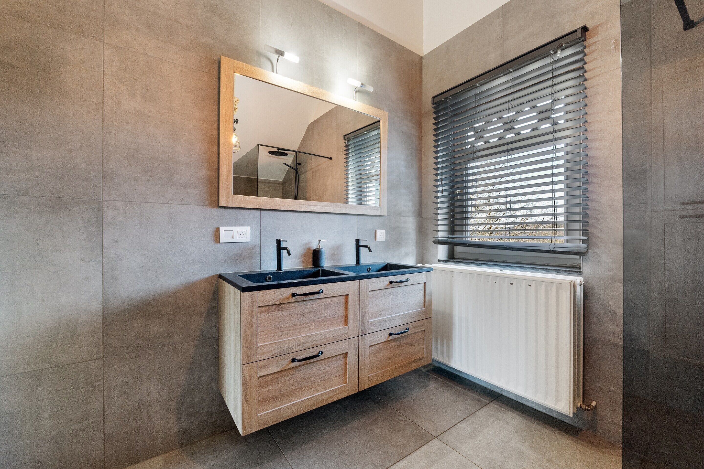 A bathroom with a double sink, wooden vanity, a mirror, and a window with blinds.