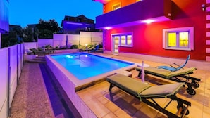 A modern outdoor pool area with a rectangular pool, lounge chairs, and a red building with illuminated windows.