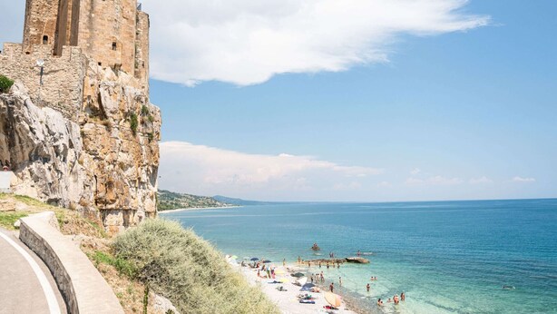 Vista costera con una torre de piedra histórica y una playa con gente y sombrillas.