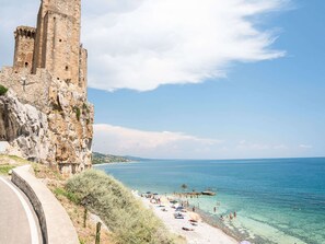 Ein Küstenblick mit einem historischen Stein-Turm und einem Strand mit Menschen und Sonnenschirmen.