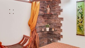 Una habitación con una silla de madera, una mesa con mantel estampado, pared de piedra, estante con adornos y un tapiz colgado.