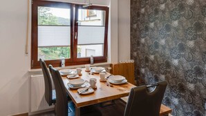 Un comedor con una mesa de madera dispuesta para cuatro personas, dos sillas, una ventana con persianas y un papel tapiz estampado.