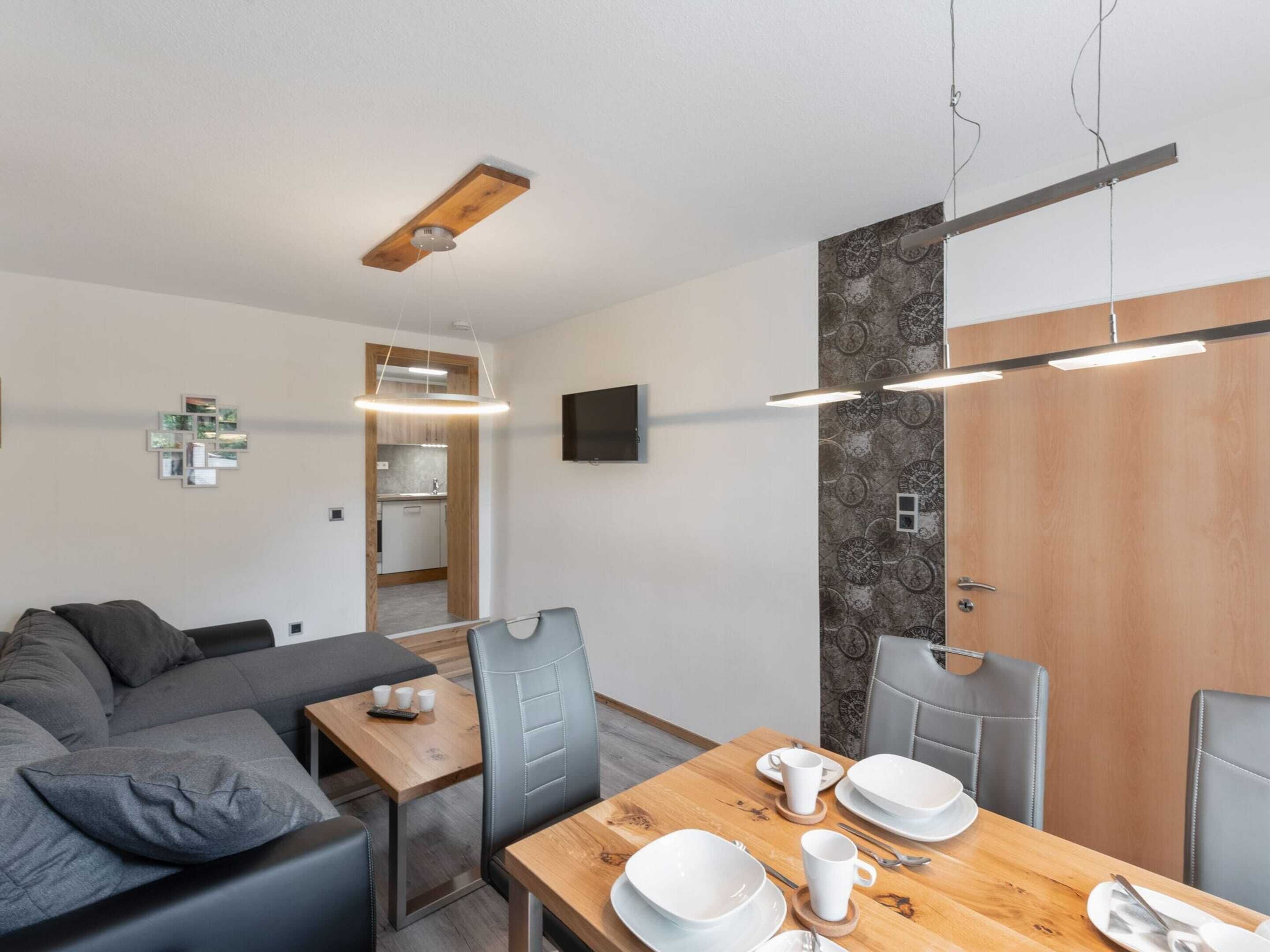 Uma sala moderna com sofá, mesa de jantar posta com pratos e xícaras, cozinha visível por uma porta aberta e uma TV montada na parede.