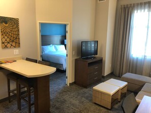 Habitación de hotel con cama, televisor sobre un mueble de madera, escritorio con silla y un pequeño rincón de descanso con otomanas.